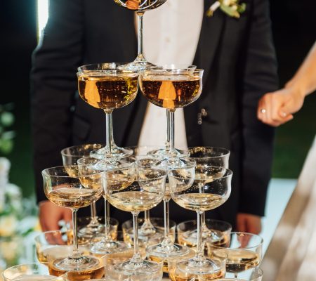 A large pyramid of champagne glasses at the evening ceremony at the arch. bride and groom are hugging A large pyramid of champagne glasses at the evening ceremony at the arch. In the background, the bride and groom are hugging.